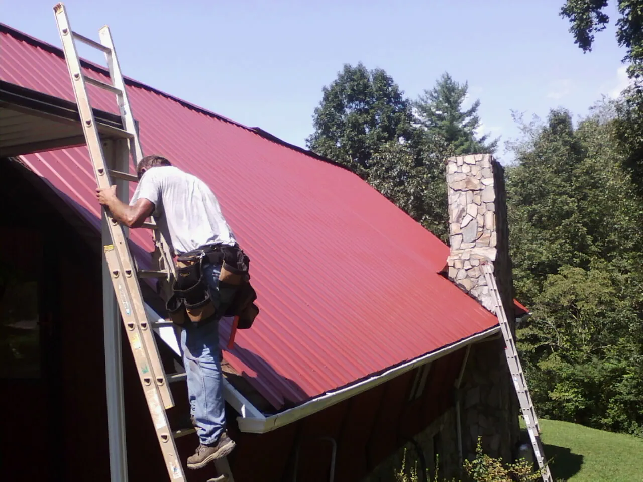 Roofer on a ladder beside a red metal roof and stone chimney — Soffit & Fascia Repair by Roofing Brothers in Bel Alton
